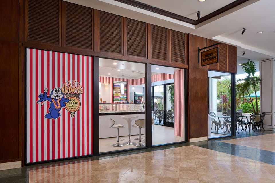 A Hawaiian-style ice cream shop with cheerful red-and-white striped décor sits in an elegant lobby, offering a sweet treat during your stay with lush outdoor seating visible beyond.