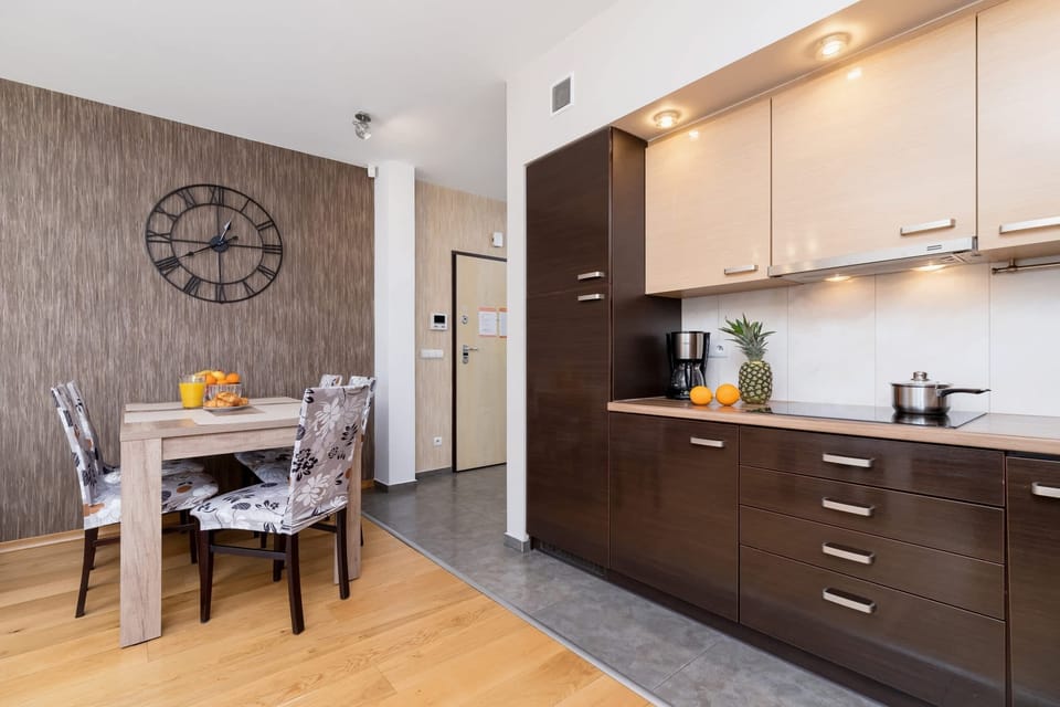 A side kitchen with brown and beige furniture is located next to the dining table above which hangs a large circular clock.
