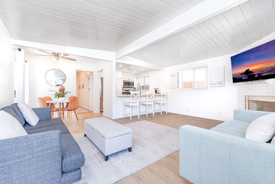 The living area includes a sofa set, streaming TV (Youtube TV), dining for four, and bar seating, all with a white, clean, beachy motif.
                