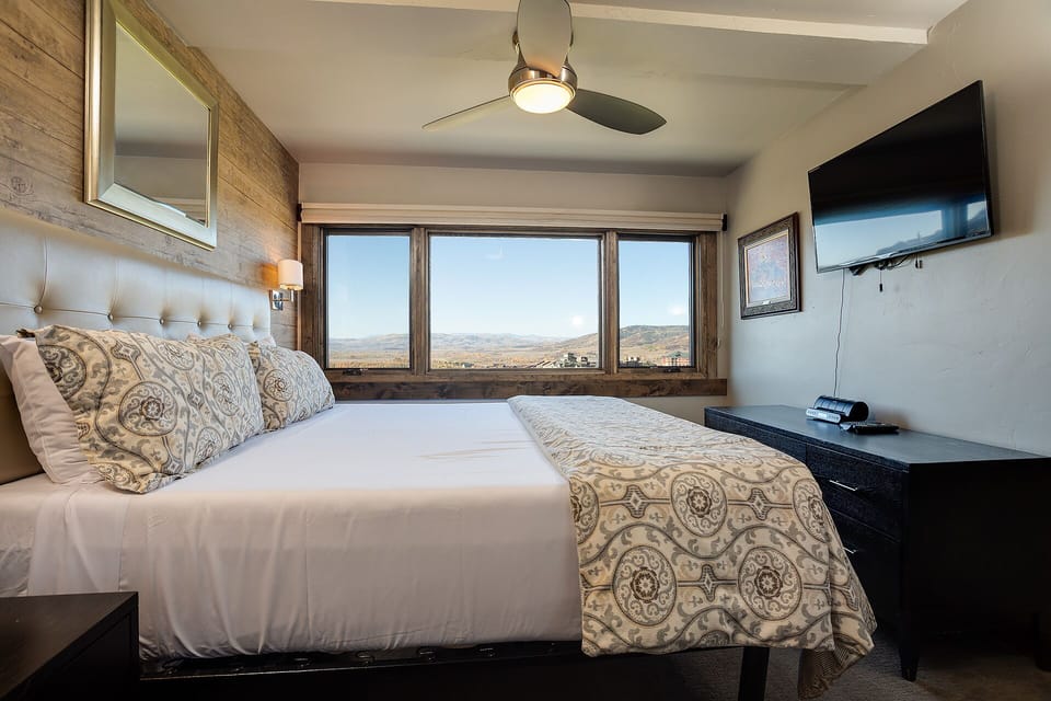 Wake up to sweeping vistas framed by rustic wood windows. Your cozy retreat features a plush tufted headboard, modern ceiling fan, and flat-screen TV for unwinding after a day exploring the landscape.