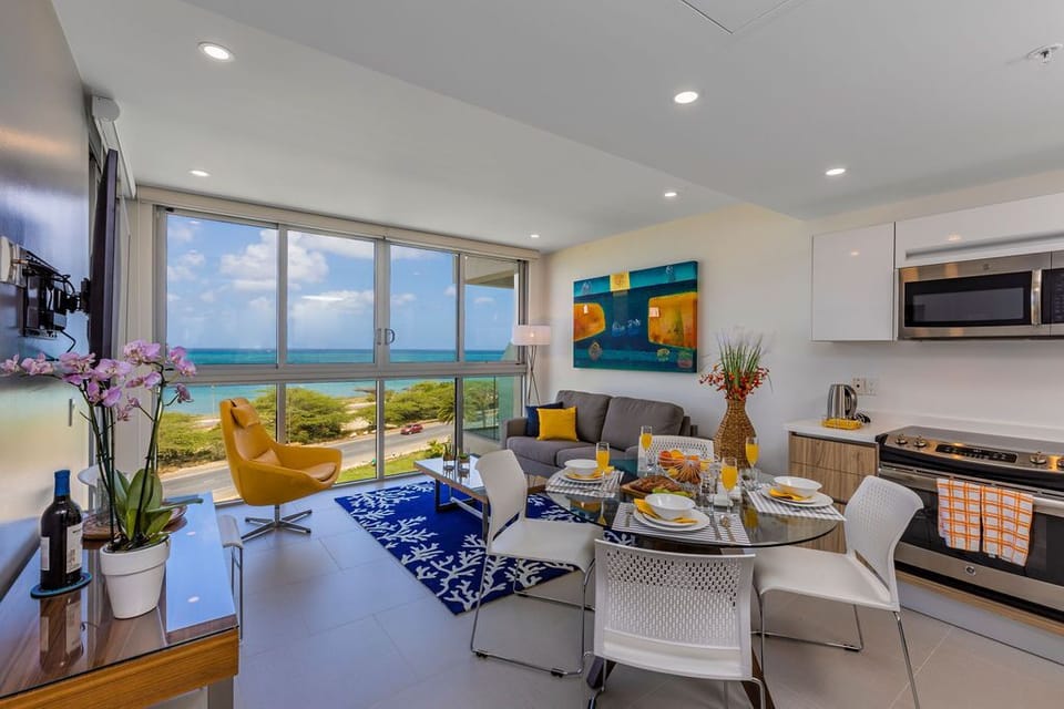 Sunlight floods your open-plan living space through floor-to-ceiling windows, framing turquoise ocean views and coastal greenery. Relax in stylish comfort where your living room, dining area, and modern kitchen blend seamlessly.