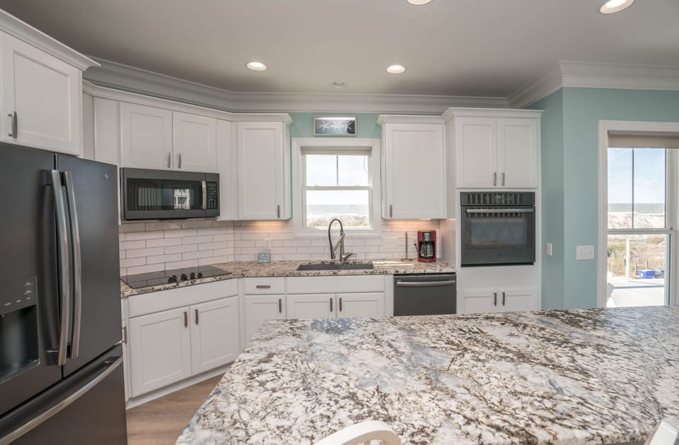 Coastal kitchen with white cabinets, granite countertops, stainless appliances, subway tile backsplash, under-cabinet lighting, and window above the sink showcasing beautiful Atlantic Ocean views in Oak Island.