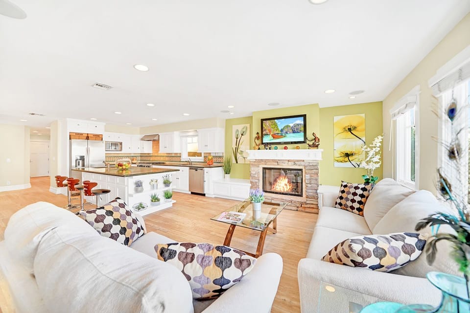 The main living area is located on the ground floor and includes two sofas, cable TV, fireplace, and kitchen and dining areas beyond.
                