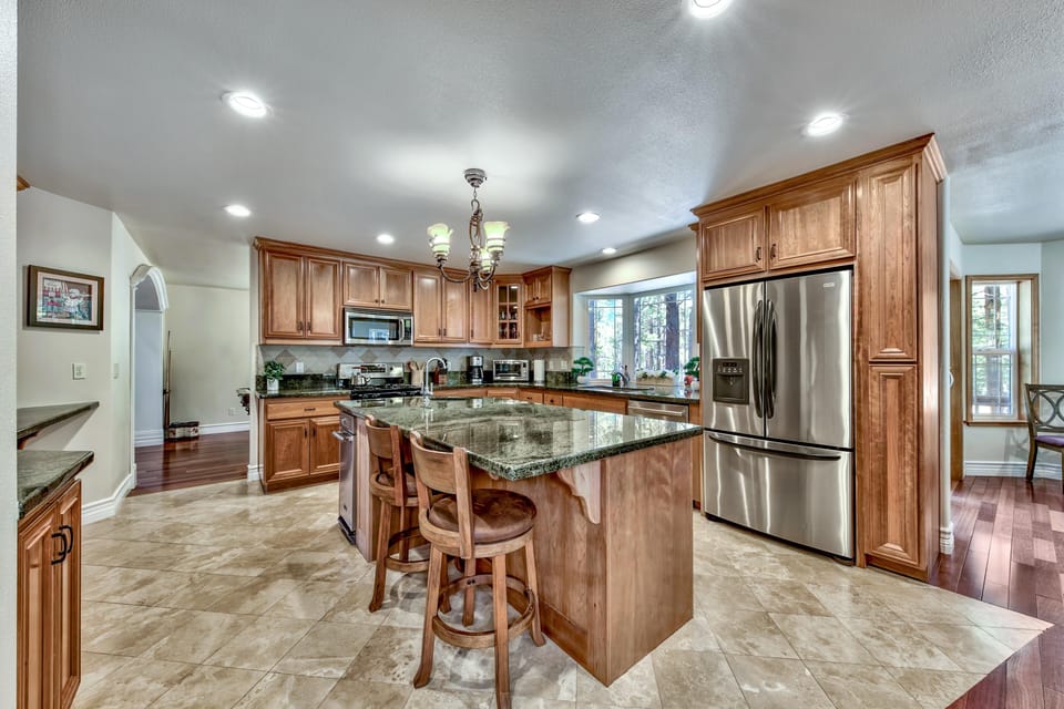 Granite-topped island with wooden stools anchors the kitchen, surrounded by honey-toned cabinetry and tiled floor.