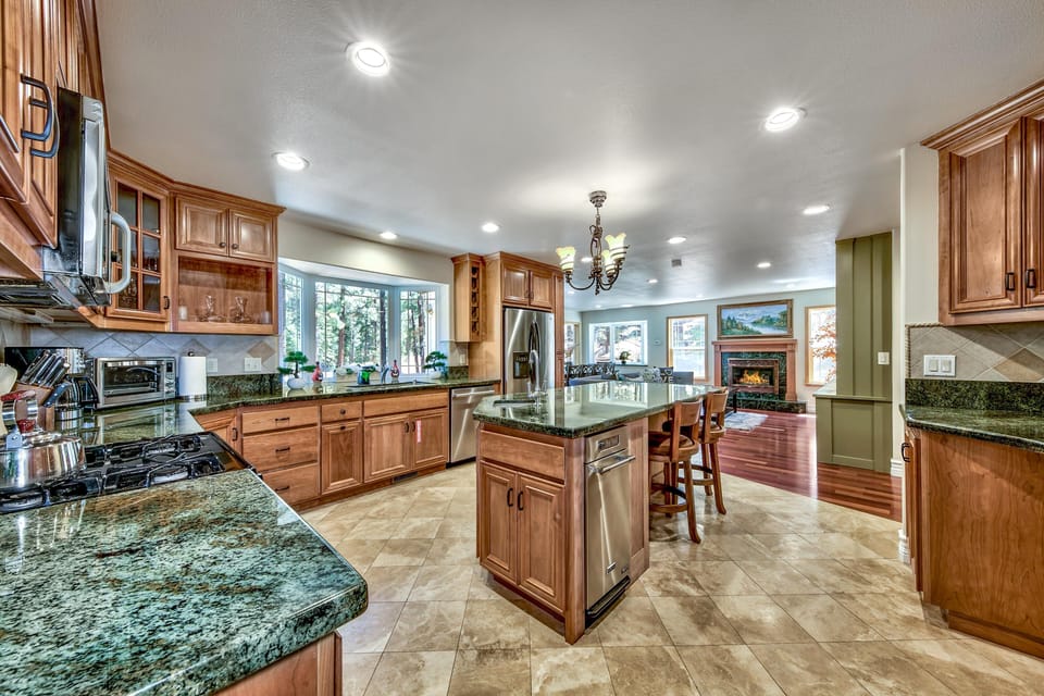 Kitchen island with granite countertop and wooden stools overlooks bay window and stainless steel appliances.