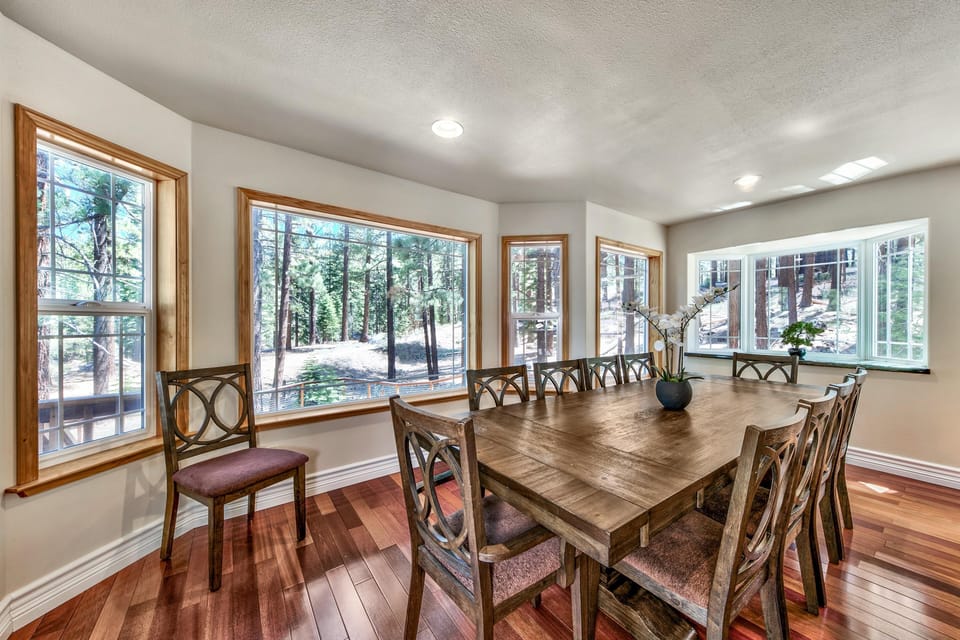 Rectangular wood dining table surrounded by ten carved wooden chairs, set beside large windows with forest views.
