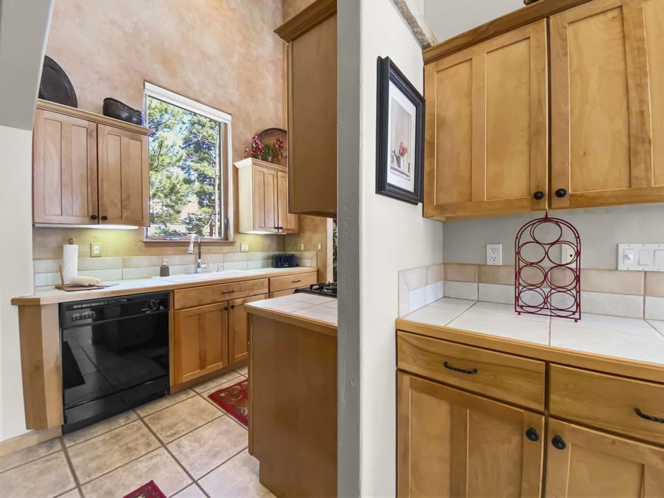 A kitchen with wooden cabinets, tiled countertops, a window above the sink, and a stove on the right. Adjacent area features additional cabinets and a decorative wine rack.