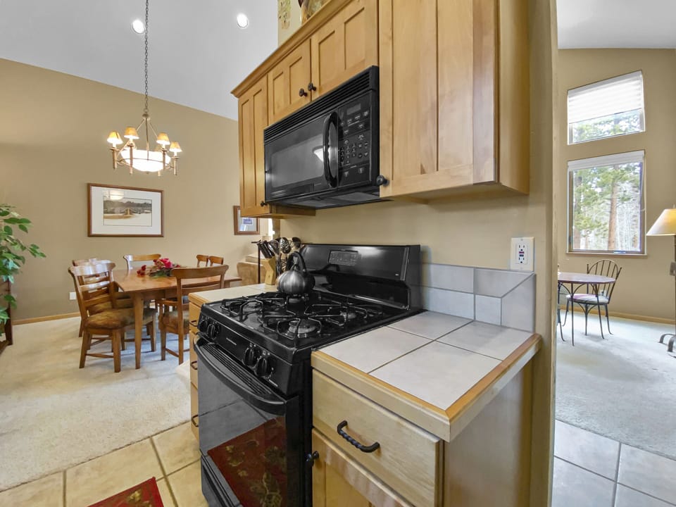 A kitchen with a black stove and microwave, wooden cabinets, a tile countertop, and a dining area with a table and chairs in the background.