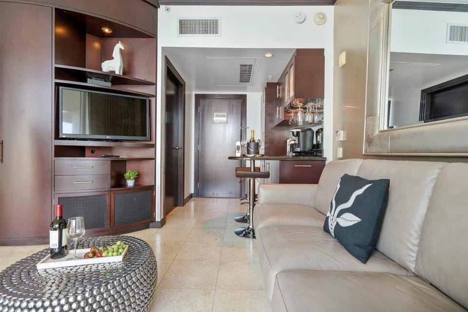 A modern hotel room with a beige sofa, a TV on a wooden cabinet, a small dining area, and a coffee station. A tray with wine, glasses, and grapes is on a metal coffee table.