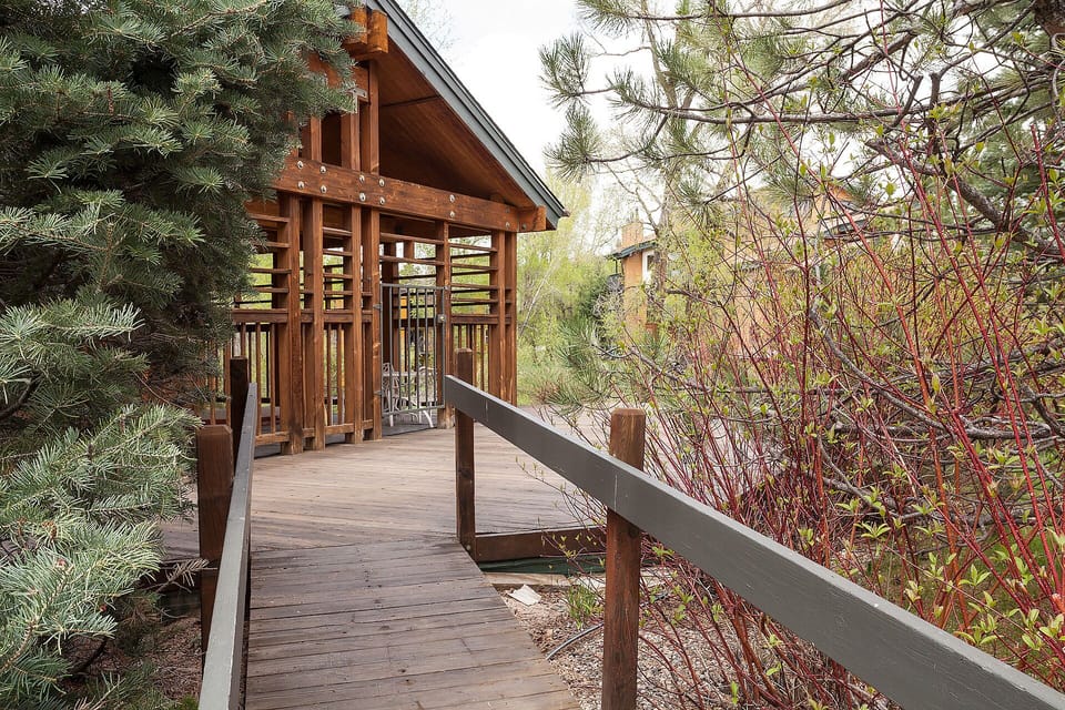 A charming wooden boardwalk leads to a cozy covered pavilion, surrounded by evergreens and seasonal foliage.