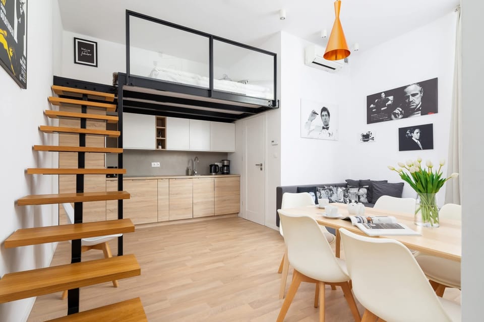 A bright space featuring a staircase leading to a loft area, with light wooden flooring and a small dining setup.