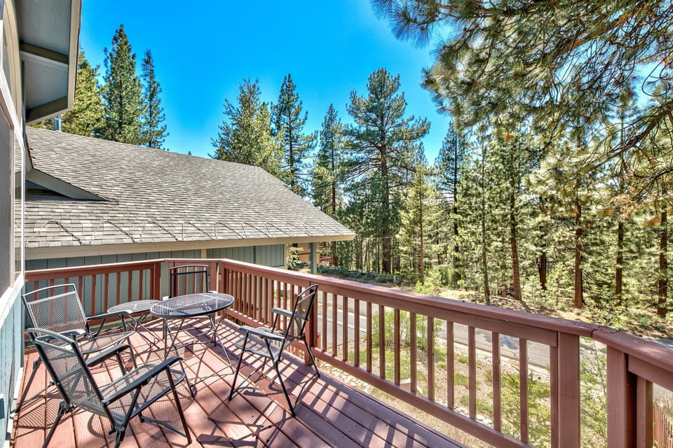 Wooden balcony with metal mesh chairs and round table overlooks tall pine trees under clear blue sky.