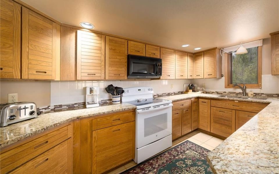 A modern kitchen with wooden cabinets, granite countertops, a white electric stove, a microwave, a toaster, and a sink under a window with natural light.