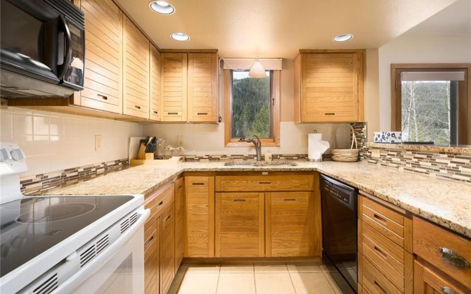 A kitchen with wooden cabinets, granite countertops, a white electric stove, and a window above the sink displaying a tree view.