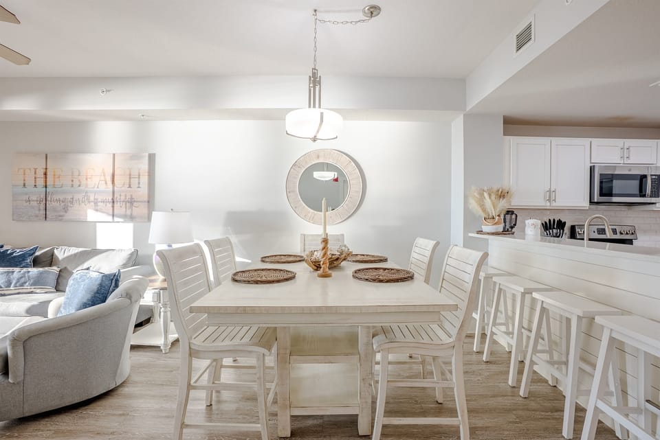 The space stays light and breezy with woven accents and a white table that somehow makes every meal feel calmer than it should. It flows right into the living room, so you can keep an eye on the kids while pretending you’re still relaxing.