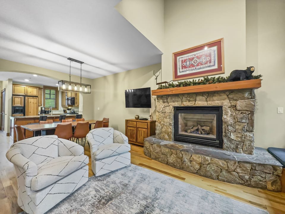 A cozy living room with a stone fireplace, two striped armchairs, and a mounted TV. The adjacent kitchen features wooden cabinets and a countertop with four bar stools. The space is adorned with warm tones.