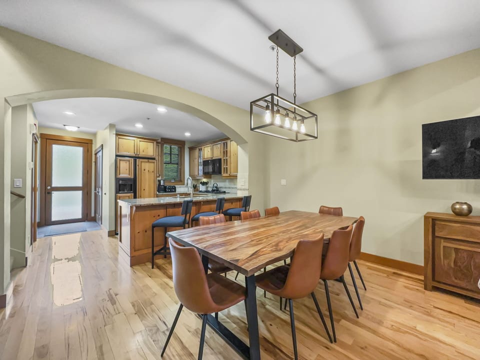 A dining room with a wooden table and six brown chairs. A kitchen with wooden cabinets is visible through an arched doorway. A pendant light hangs above the table, and a sideboard is against the wall.