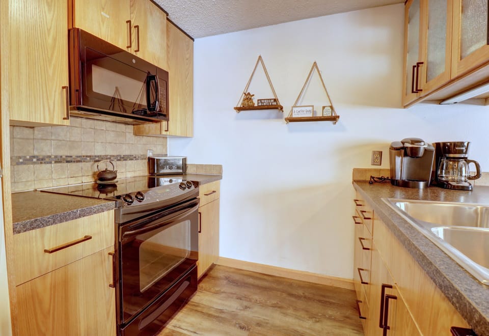 A small kitchen with wooden cabinets, black appliances, a microwave, a coffee maker, a toaster, and hanging triangle-shaped shelves on a white wall.