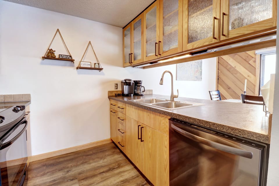 A modern kitchen with wooden cabinets, a double sink, a dishwasher, and a coffee maker. Two triangular shelves are mounted on the wall, and a stove with an oven is on the left.