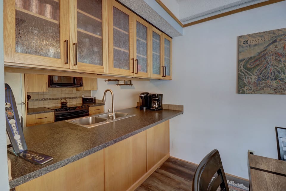 A small kitchen with wooden cabinets, a double sink, a microwave, a coffee maker, and a ski leaning against the counter. There's a map artwork on the wall.