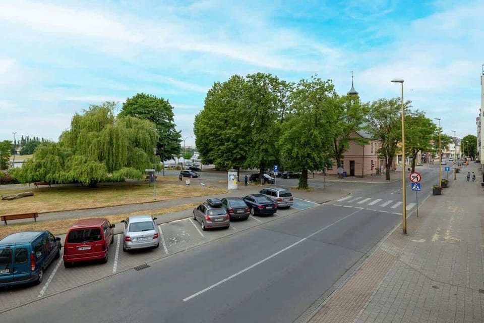 Widok na ulicę z zaparkowanymi samochodami i zielony skwer po drugiej stronie ulicy.