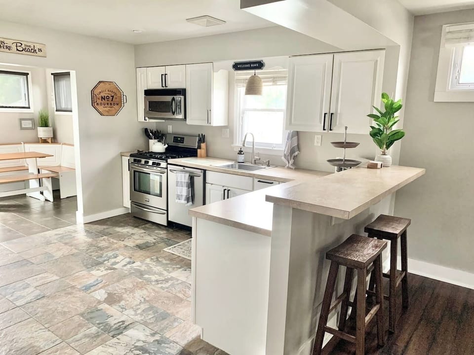 This modern kitchen features stainless steel appliances, a deep sink, and a raised breakfast bar with wooden stools. The open layout leads directly into a sunlit corner breakfast nook, providing a cozy spot for morning coffee.