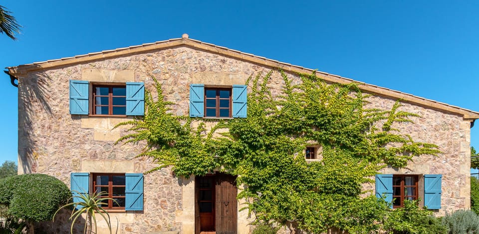 Traditional Mallorcan stone facade with blue shutters and greenery, offering rustic Mediterranean charm.