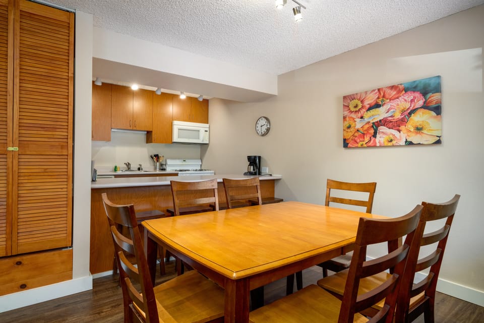 A cozy kitchen and dining area featuring a wooden table with six chairs, wooden cabinets, a wall clock, a floral painting, a microwave, and a coffee maker.