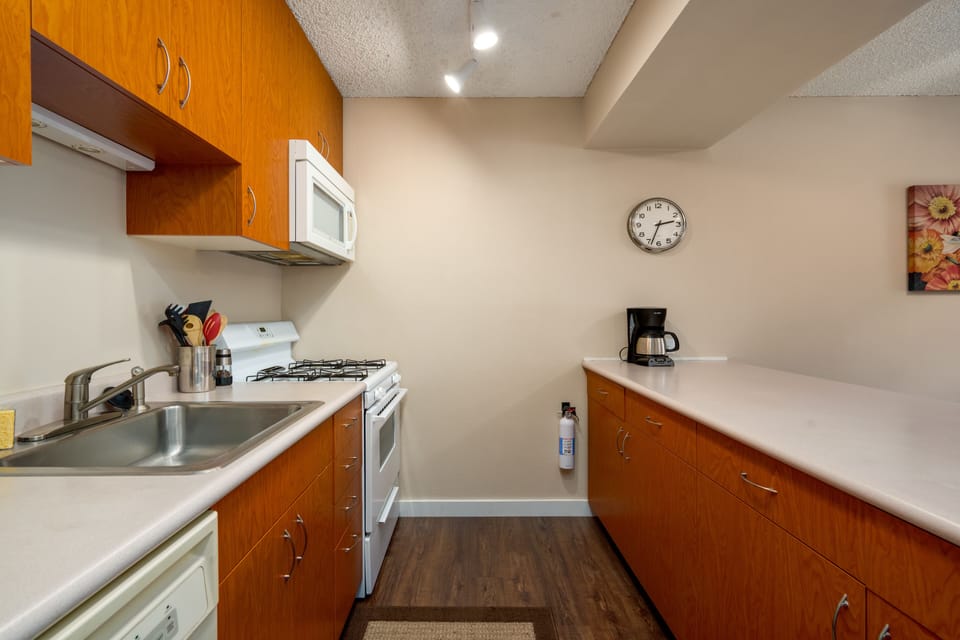 Kitchen with wooden cabinets, white countertops, sink, stove, white microwave, coffee maker, wall clock, and floral wall art.