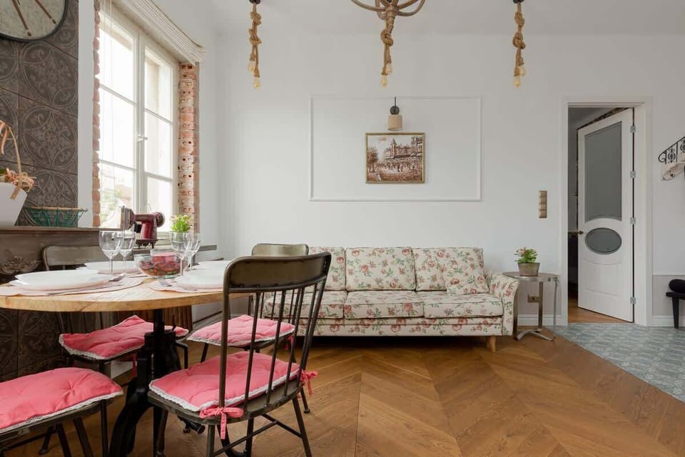 A bright dining area with vintage-inspired decor, a sturdy wooden table, floral-patterned chairs, and large windows for a homely ambiance.