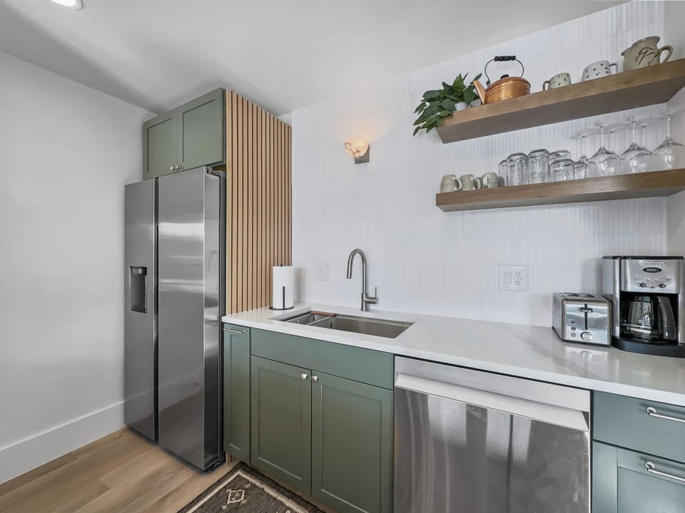 Modern kitchen with green lower cabinets, stainless steel appliances, open shelving, a sink beneath a wall sconce, countertop appliances, and a wooden slat accent wall beside a double-door refrigerator.