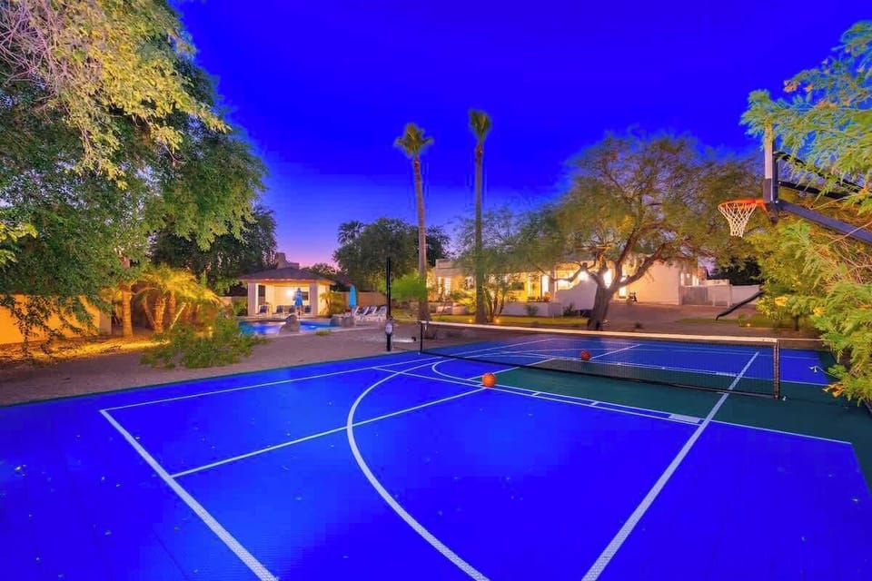 A striking twilight view captures the property's multi-sport court, surrounded by palm trees, lush landscaping, and warm outdoor lighting that illuminates the entire compound.