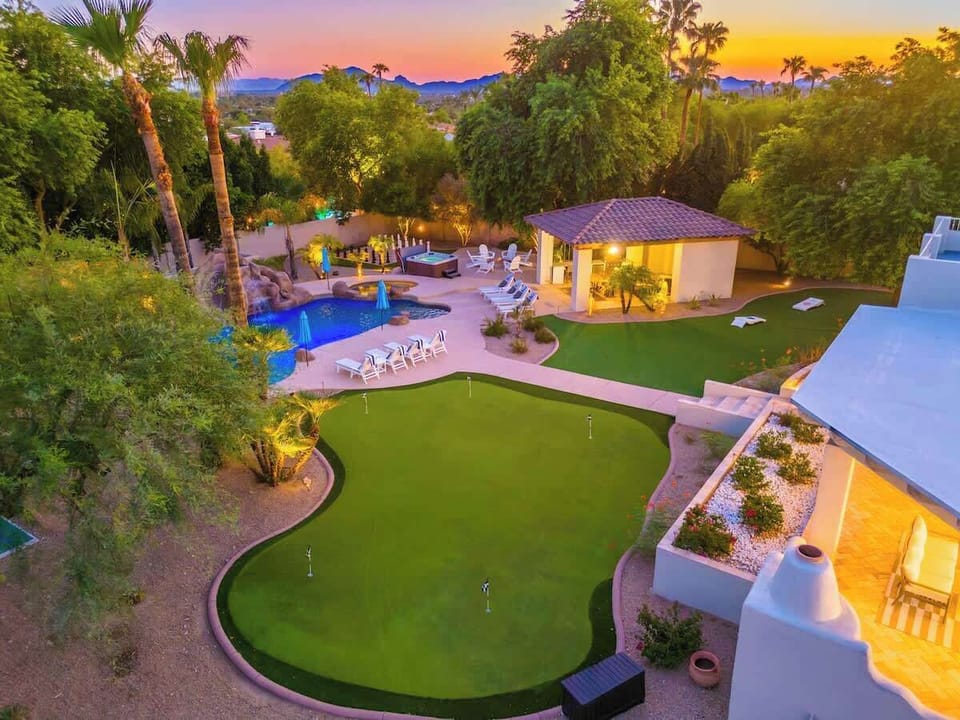 Aerial view of a private desert retreat showcasing a putting green, sparkling pool, cabana, and lush palm-lined grounds at dusk with mountain backdrop.