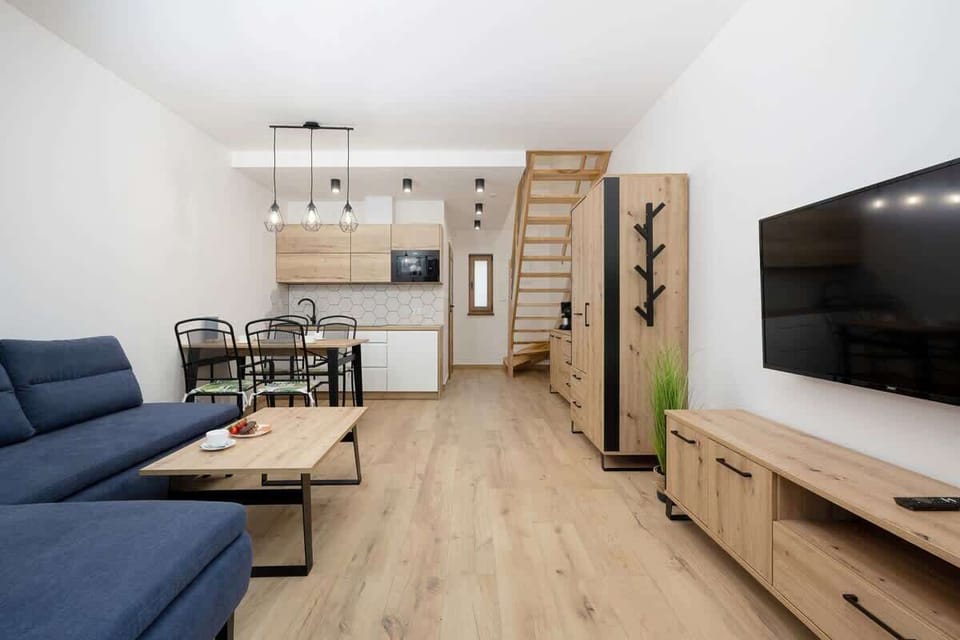 Another angle of the living room showing the dining area in the background. The layout is open-plan, with a small dining table and chairs near the back, maintaining a natural, wooden aesthetic throughout.