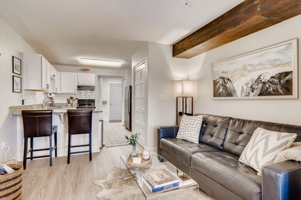 A cozy living room with a black leather couch, a glass coffee table, and a kitchen in the background. Kitchen has white cabinets, black appliances, and a small breakfast bar with two stools.