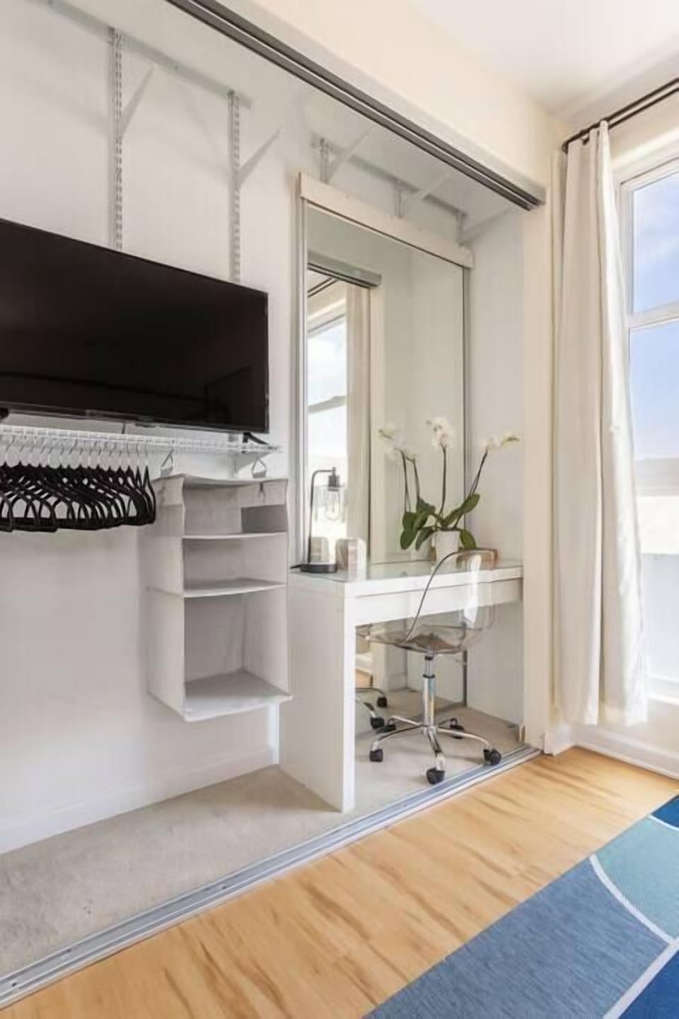 Prepare for your day at this bright, minimalist vanity featuring a sleek white desk, a ghost chair, and a large mirror that reflects the natural light of the room.