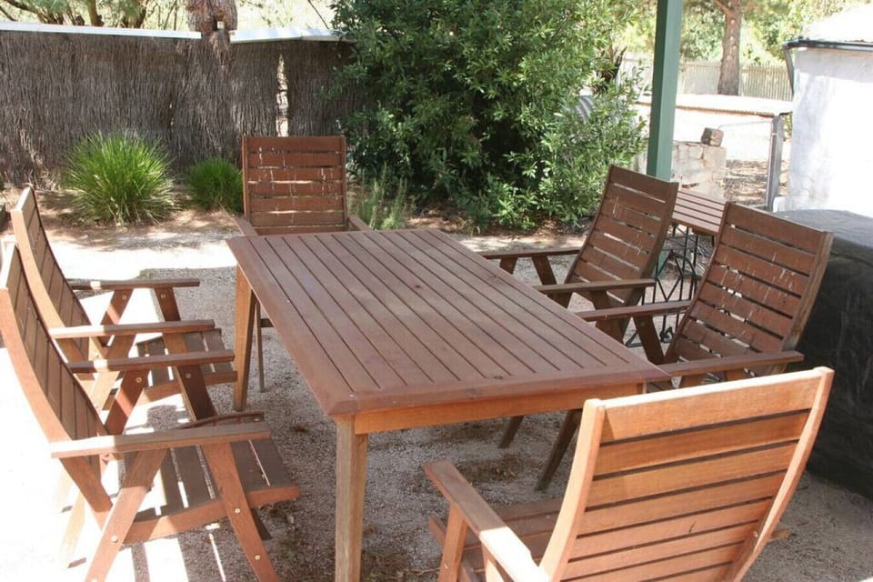 This outdoor setting features a sturdy wooden dining table surrounded by six matching chairs, perfect for enjoying meals or gatherings in a shaded garden area. The space is framed by lush greenery and a natural privacy screen, creating a relaxed and invit