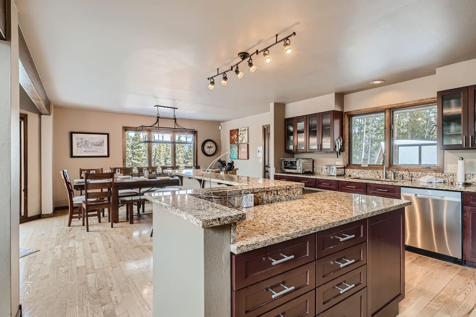 Modern kitchen with granite countertops, wooden cabinets, an island, and stainless steel appliances; adjacent dining area with a wooden table and large windows providing natural light.