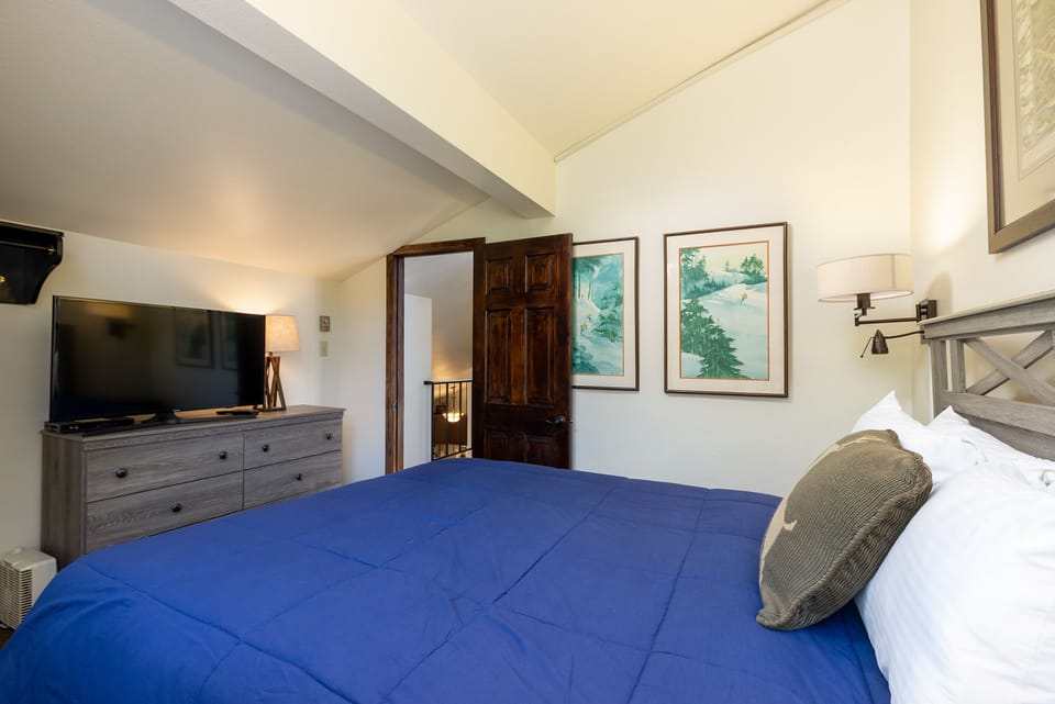 A bedroom with a wooden dresser and a TV, a bed with blue bedding, two wall art pieces, and an open wooden door leading to a hallway.