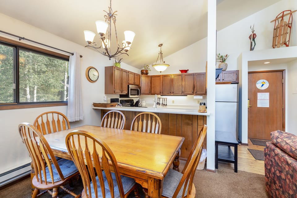 A kitchen and dining area with wooden cabinets, a wooden dining table and chairs, stainless steel appliances, chandeliers, and a window with a wooded view.