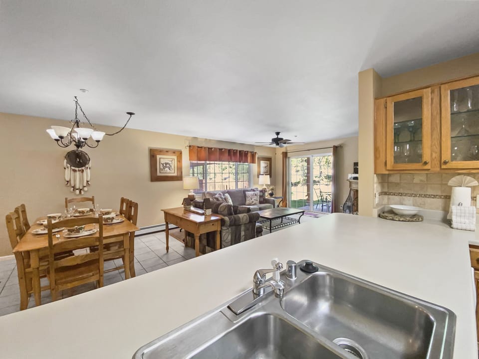 A bright and spacious open-plan kitchen and living area, featuring wooden dining furniture, a beige sofa set, large windows with sunlight streaming in, and light-colored tiles on the floor.