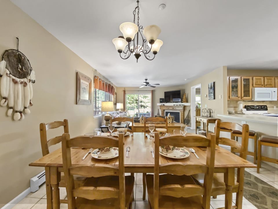 A dining area with a wooden table and chairs set for four, surrounded by a kitchen and living room in an open floor plan. A chandelier hangs above the table, and a window offers a view outside.