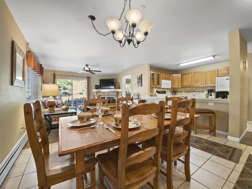 A dining area with a wooden table and eight chairs, set with plates and glasses. The background shows a living area with a couch, a TV, and a kitchen with wooden cabinets and appliances.