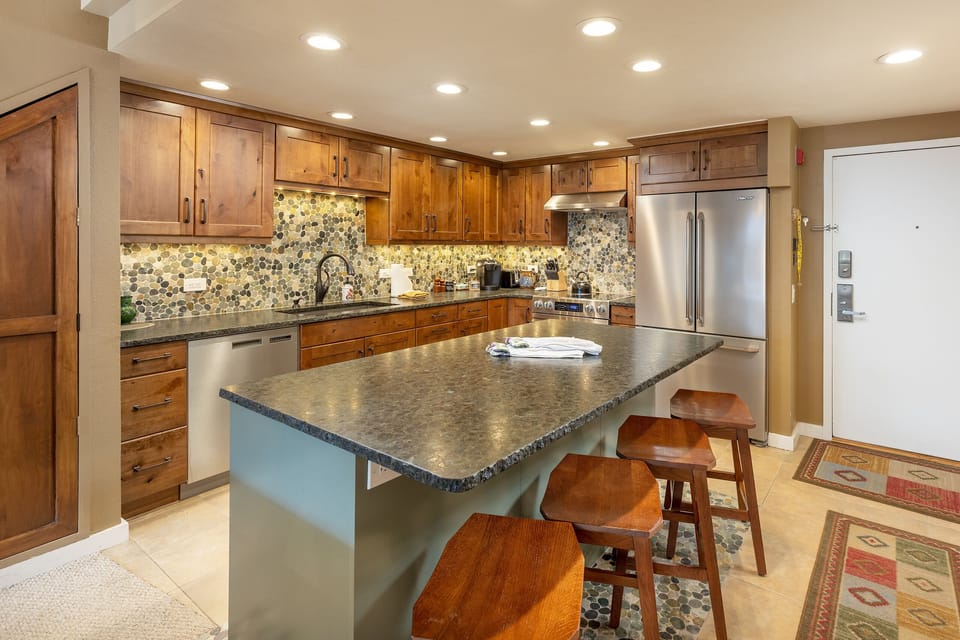 Modern kitchen featuring a central island with four wooden stools, wooden cabinets, a tiled backsplash, a stainless steel refrigerator, and various kitchen appliances.