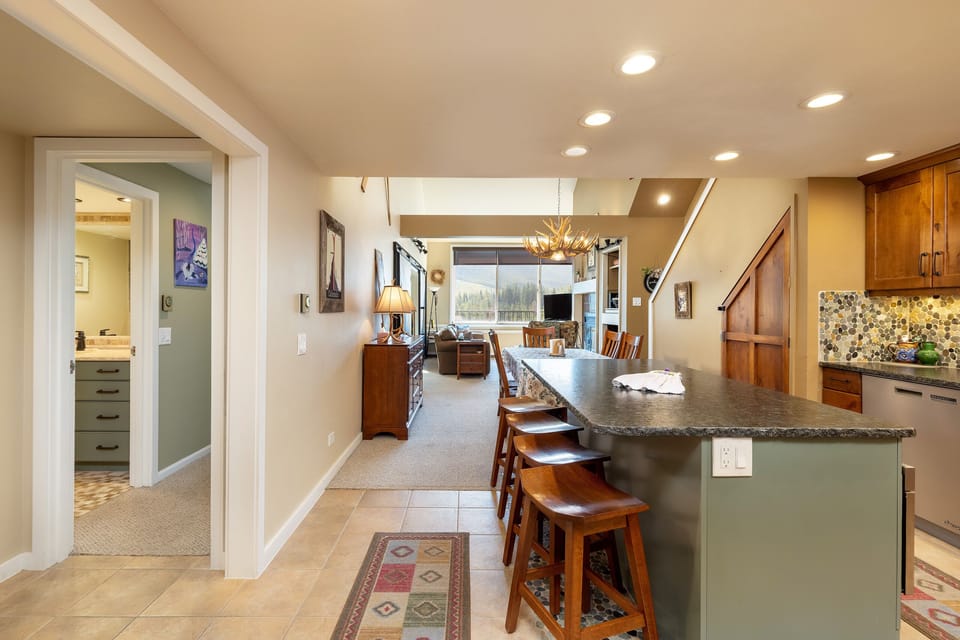 Open-concept kitchen and living area with an island and several stools in the foreground. The living area has a large window with a scenic view, and there are doors leading to other rooms.
