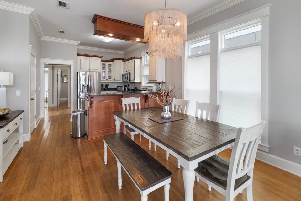 Gather around the rustic farmhouse table under a statement chandelier, where natural light pours through tall windows and modern appliances meet warm wood cabinetry in this inviting open-concept space.