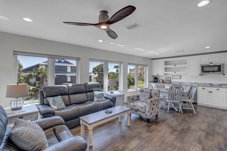 - Bright living space with plush leather recliners and large windows all around
- Open concept layout flows into dining nook and sleek white kitchen area
- Palm views, natural light, and warm floors make this the perfect gathering spot