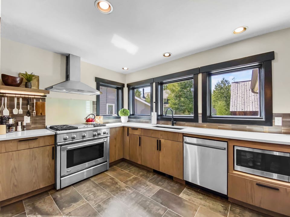 Modern kitchen with stainless steel appliances, wooden cabinets, extensive countertop space, large windows for natural light, and tiled flooring.