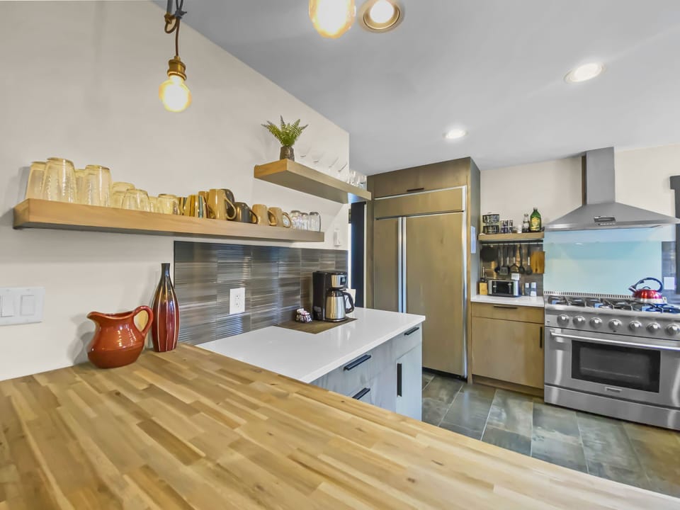 Modern kitchen with stainless steel appliances, wooden countertops, floating shelves with mugs and glasses, a potted plant, and pendant lighting.