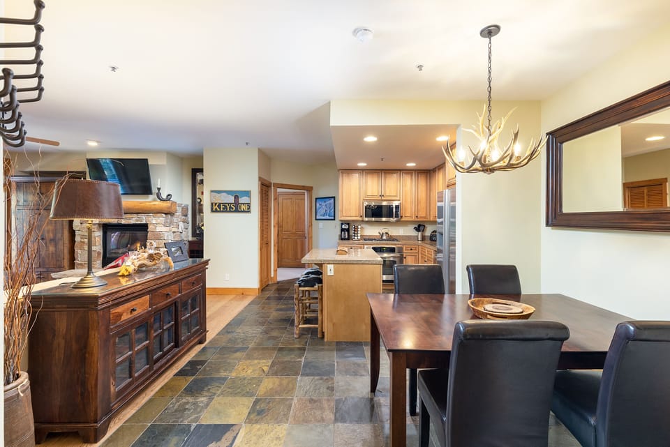 A kitchen and dining area with wooden furniture, a kitchen island, a mounted TV, a rustic chandelier, and a large mirror on the wall, featuring stone flooring and neutral-colored walls.